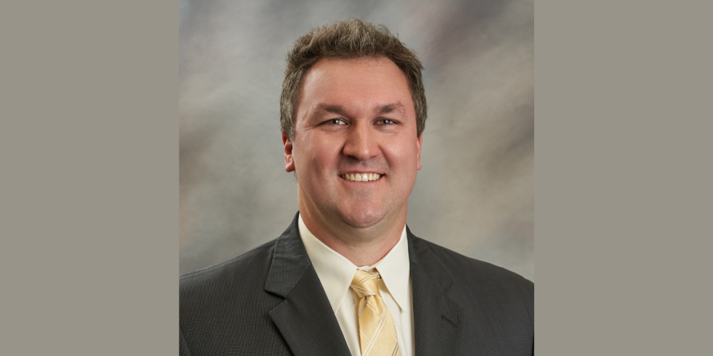 Professional headshot of Jason Wright in a dark suit, cream shirt, and gold tie against a neutral studio background.