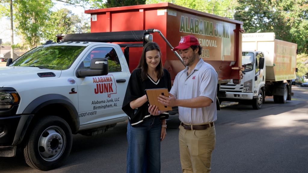 Chase Neblett (right) wearing a red signature hat of AL Junk Removal with a white shirt, showing a clipboard to a dark haired young woman in black shirt and blue jeans in front of an AL Junk dumpster truck.