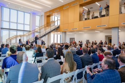 A large group of people attend an event in a modern, brightly lit auditorium with a speaker at a podium and attendees seated in rows.